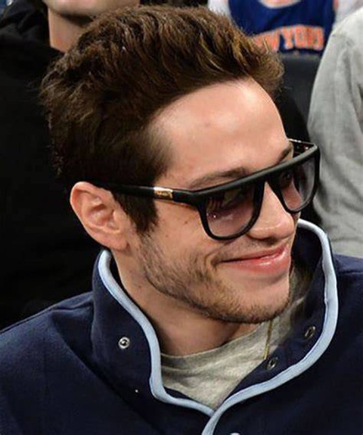 Pete Davidson is wearing sunglasses and a hat that says "Freak'n Easy" on a red carpet. 